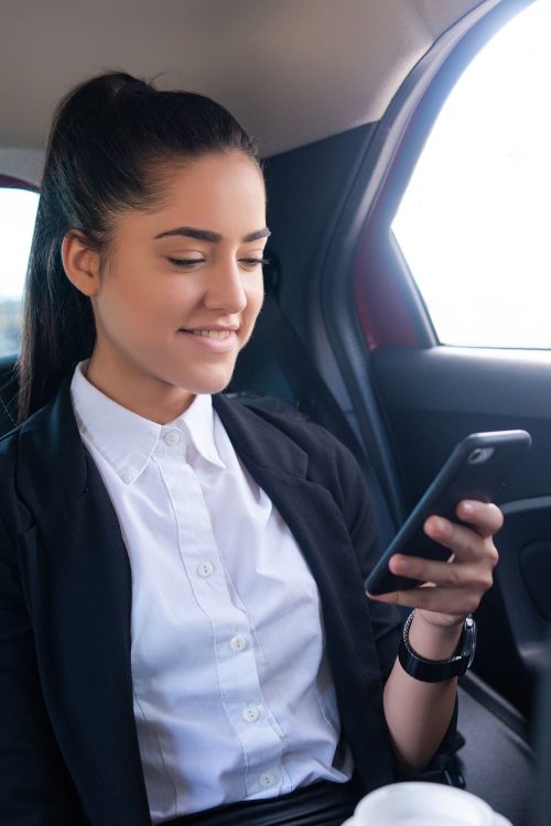 A woman in a business suit using her cell phone in the back seat of a car.