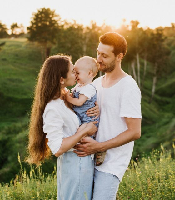 Ein Paar küsst sein Baby bei Sonnenuntergang auf einem Feld.